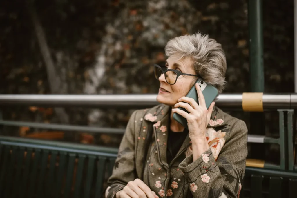 Elderly woman with glasses talking on a smartphone while seated outdoors.